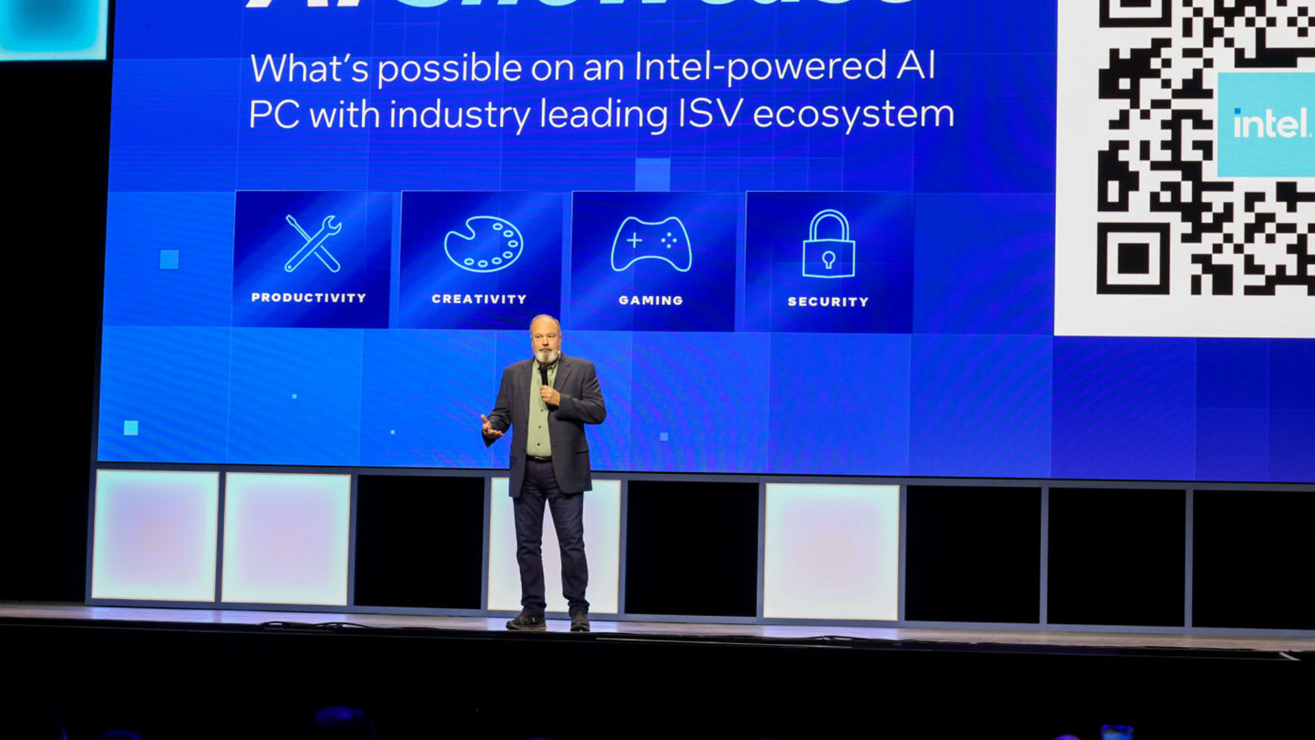 A speaker stands on stage in front of a large screen displaying AI Showcase with icons labeled Productivity, Creativity, Gaming, and Security. A QR code and an Intel logo are also visible on the screen.