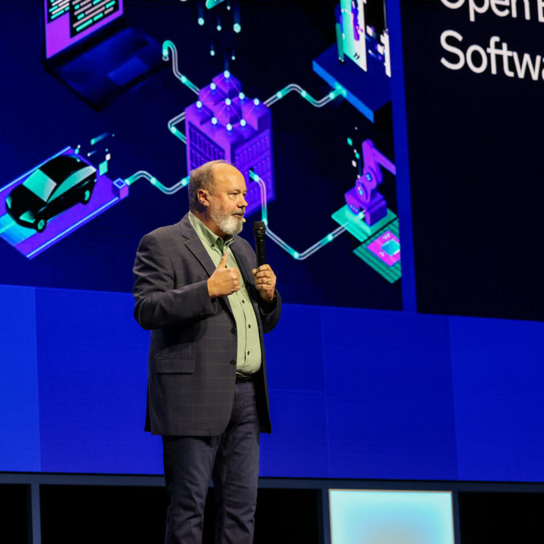 A person speaks on a stage in front of a digital screen showing futuristic graphics and the words Open Edge Software. They wear a suit and stand against a backdrop with shades of blue and purple.