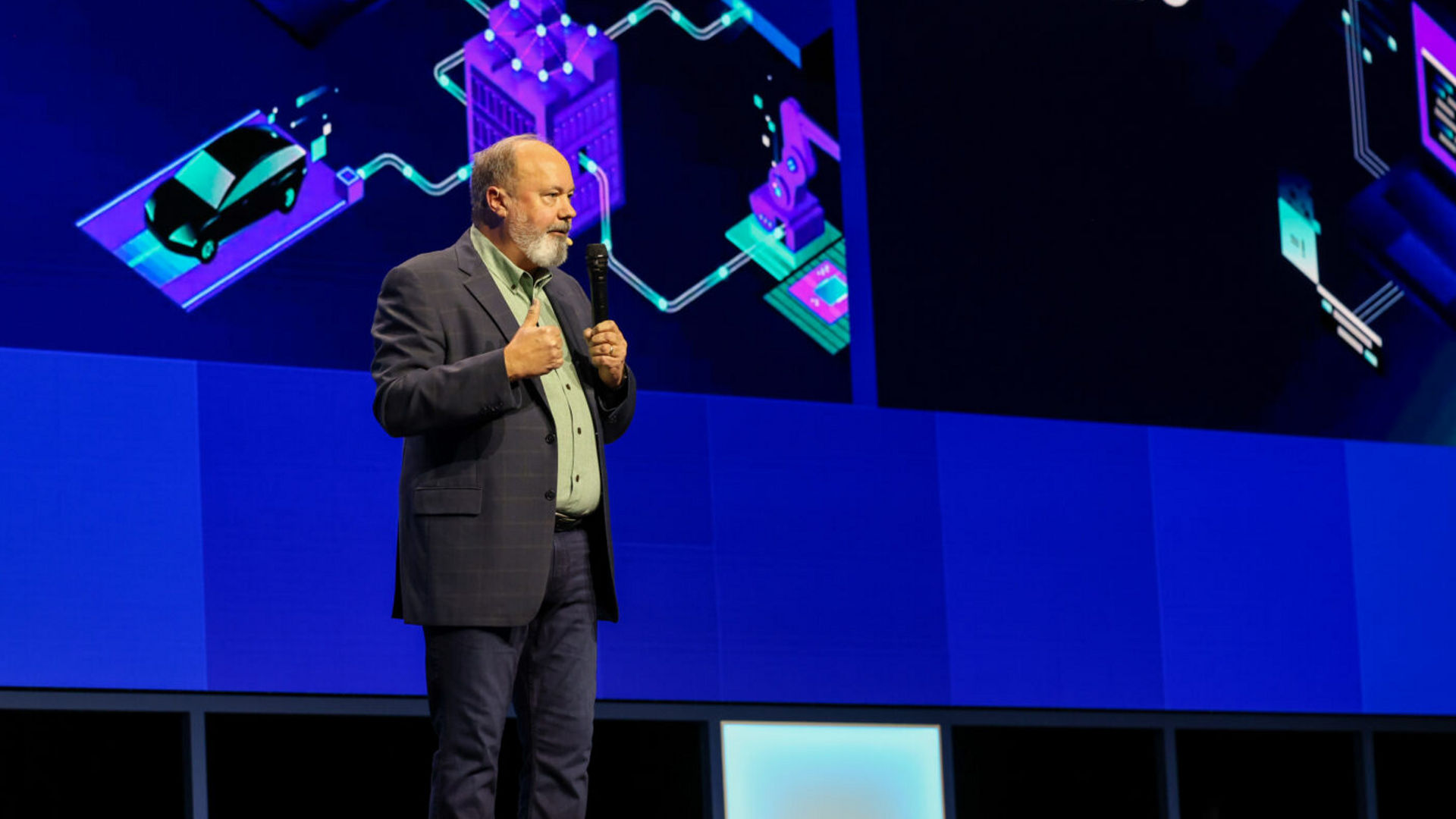 A person speaks on a stage in front of a digital screen showing futuristic graphics and the words Open Edge Software. They wear a suit and stand against a backdrop with shades of blue and purple.