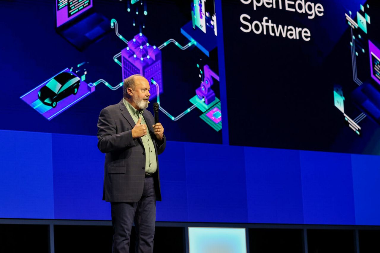 A person speaks on a stage in front of a digital screen showing futuristic graphics and the words Open Edge Software. They wear a suit and stand against a backdrop with shades of blue and purple.