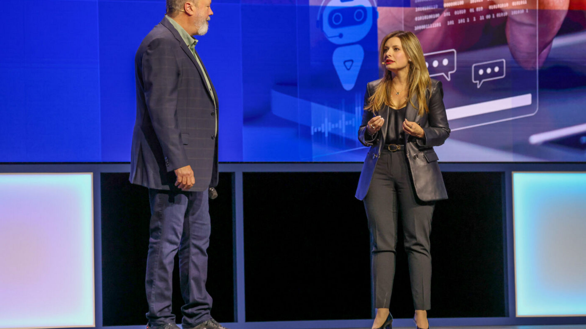 A man and woman are standing on a stage in front of a large screen with digital graphics showing AI technology. They appear to be engaged in conversation during a presentation or conference.