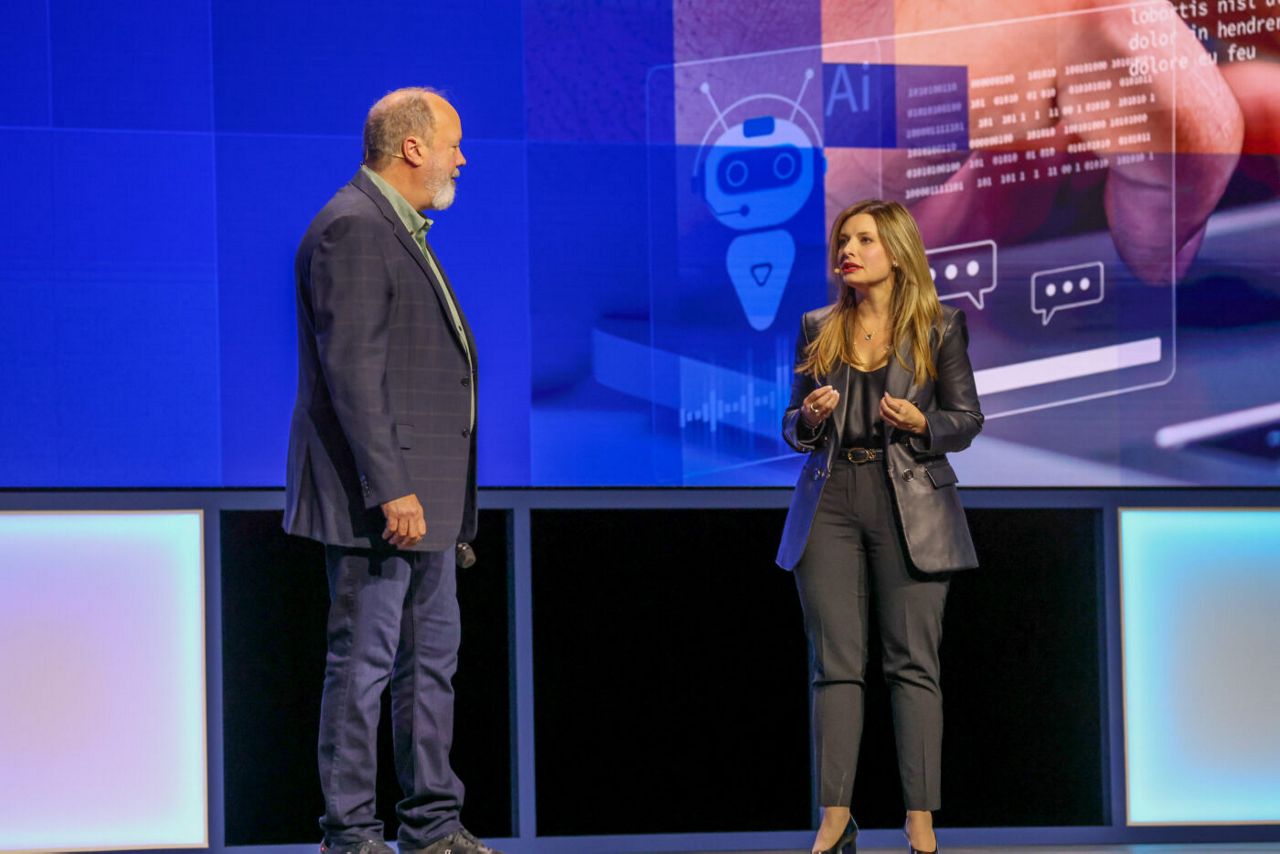 A man and woman are standing on a stage in front of a large screen with digital graphics showing AI technology. They appear to be engaged in conversation during a presentation or conference.
