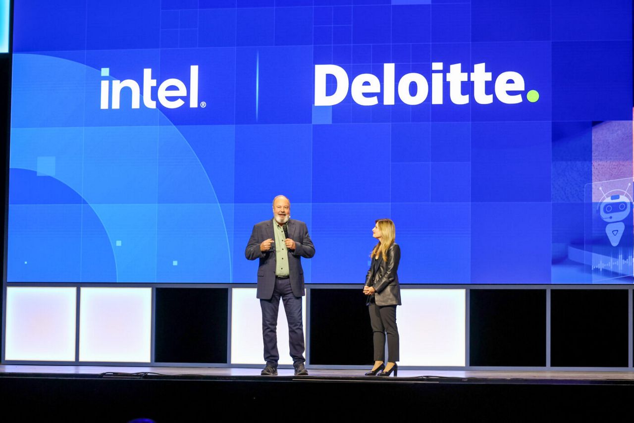 Two people stand on stage with a blue background displaying the logos for Intel and Deloitte. The man is speaking into a microphone while the woman listens attentively. The setting appears to be a professional conference or event.