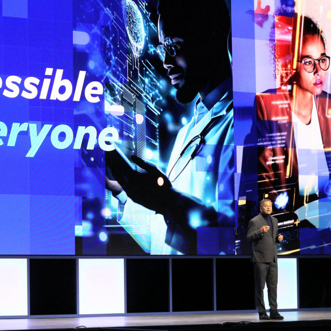 A speaker stands on a stage in front of a large screen displaying the text AI accessible to everyone. The screen shows digital imagery including a person interacting with a holographic interface, symbolizing technology and artificial intelligence.