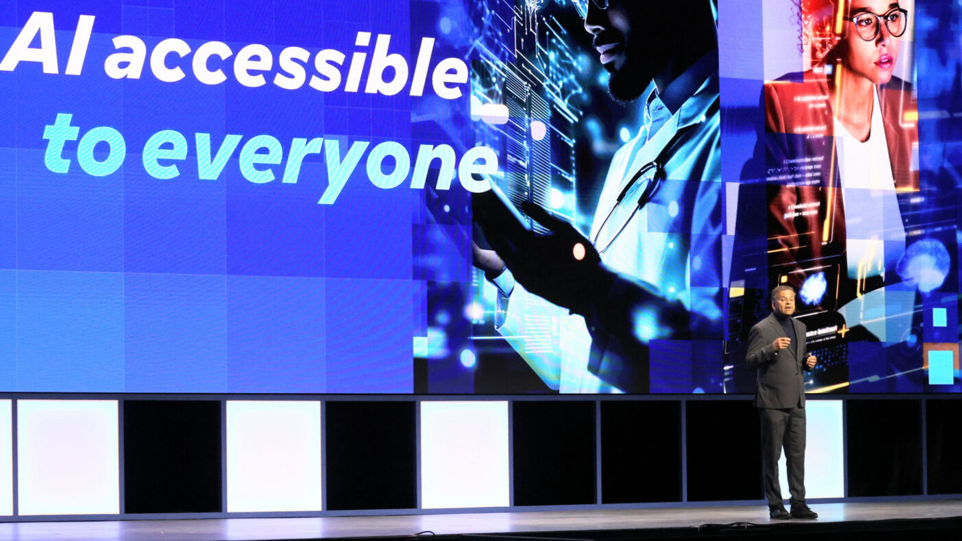 A speaker stands on a stage in front of a large screen displaying the text AI accessible to everyone. The screen shows digital imagery including a person interacting with a holographic interface, symbolizing technology and artificial intelligence.