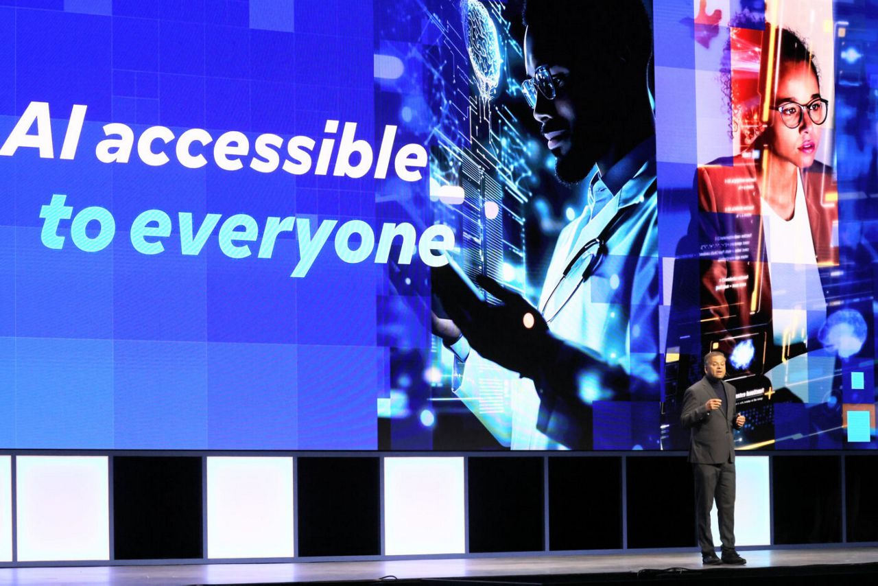 A speaker stands on a stage in front of a large screen displaying the text AI accessible to everyone. The screen shows digital imagery including a person interacting with a holographic interface, symbolizing technology and artificial intelligence.
