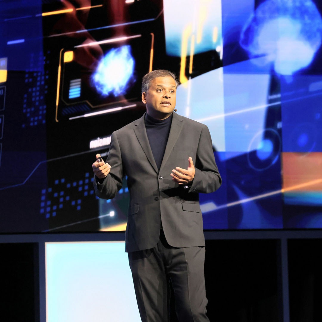 A person in a dark suit with a black turtleneck is speaking on stage. They are holding a presentation device. Behind them, there is a digital screen displaying abstract blue and orange graphics.