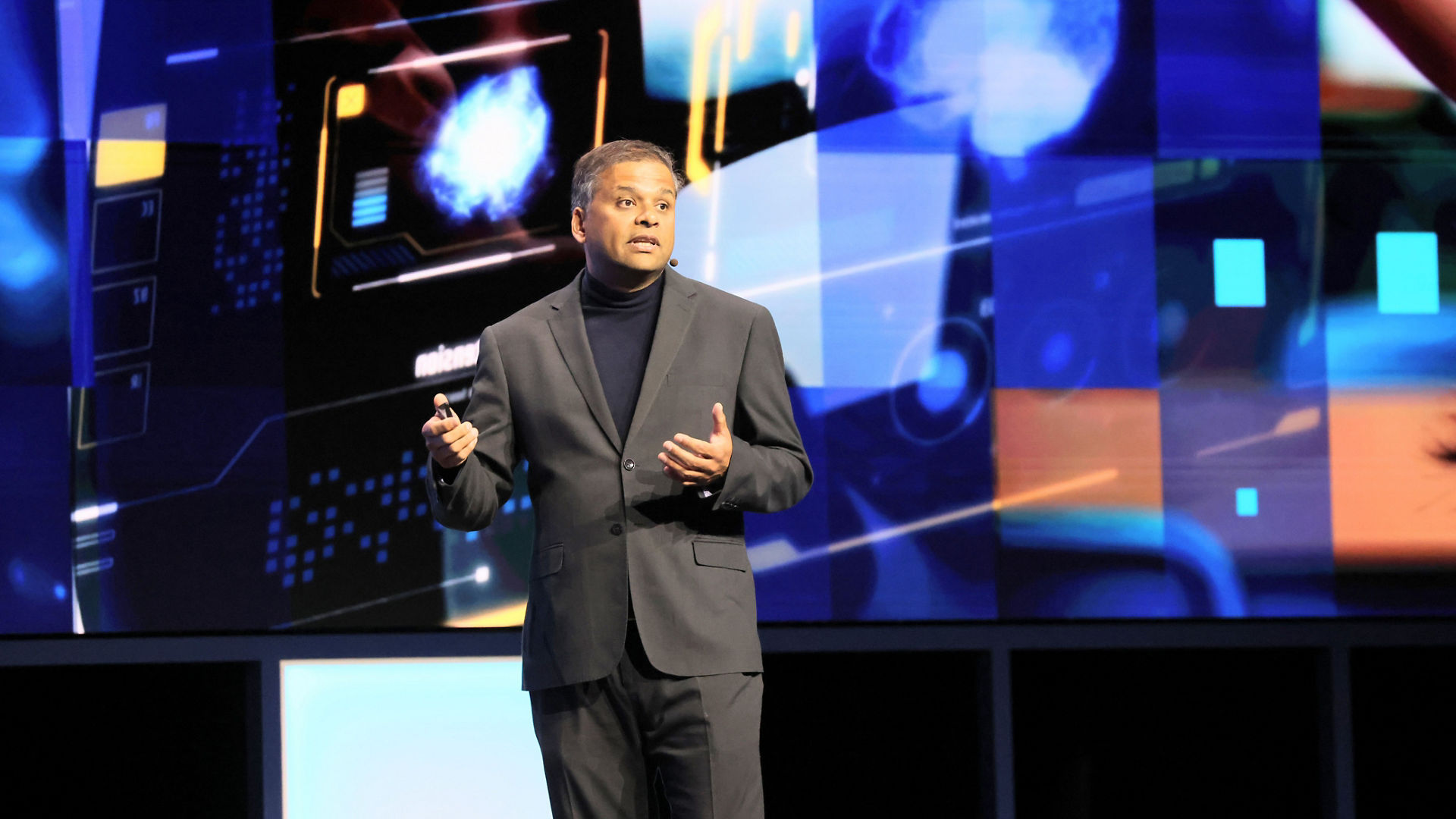 A person in a dark suit with a black turtleneck is speaking on stage. They are holding a presentation device. Behind them, there is a digital screen displaying abstract blue and orange graphics.