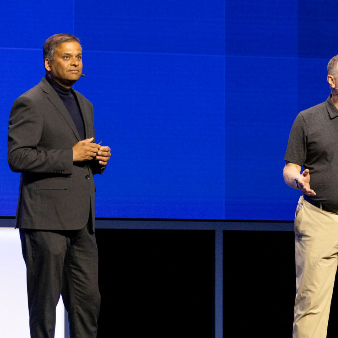 Two men are standing on stage speaking. The man on the left is wearing a dark suit, while the man on the right is in a black polo shirt and khaki pants. They appear to be engaged in a discussion or presentation against a blue geometric background.