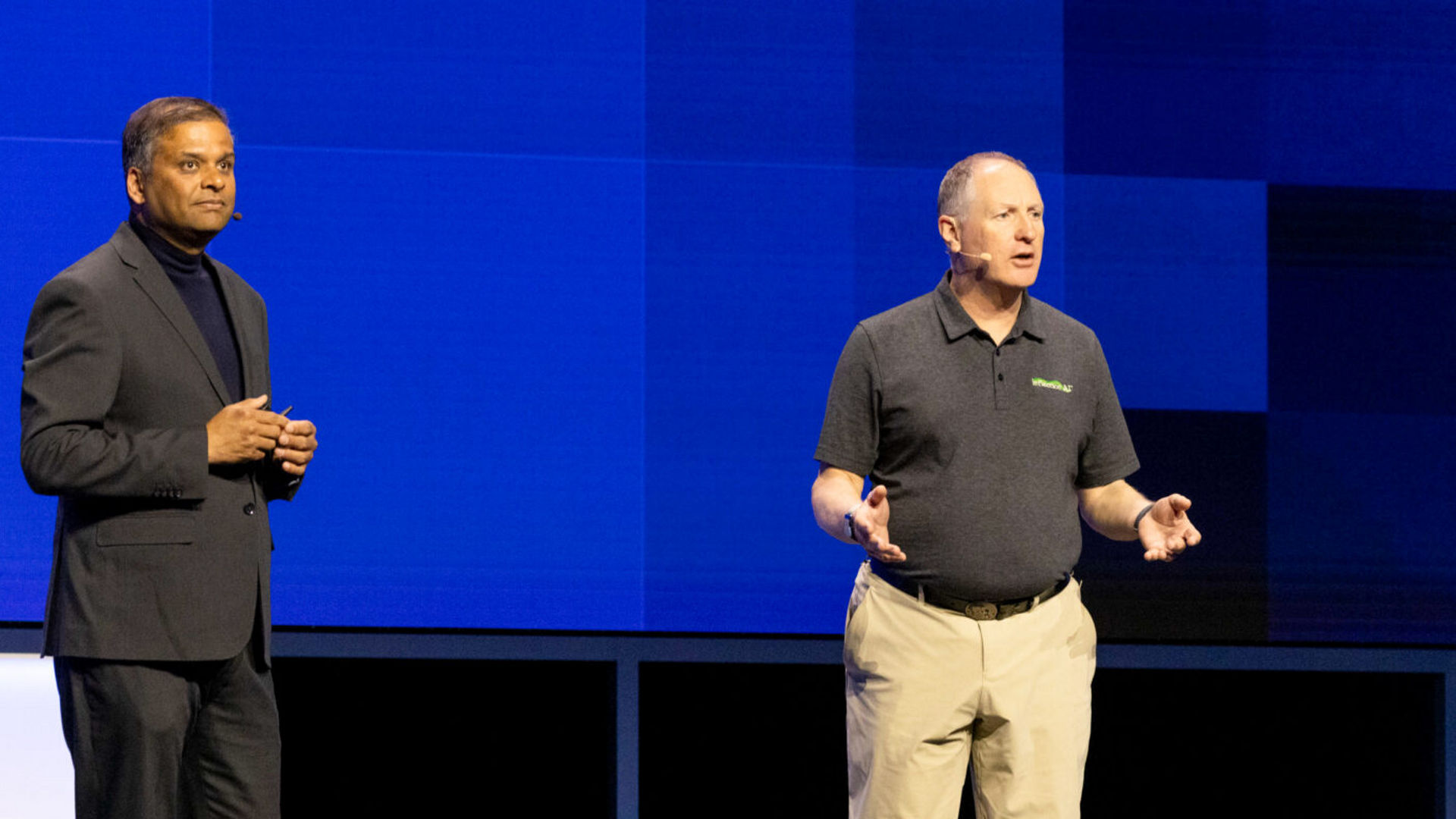 Two men are standing on stage speaking. The man on the left is wearing a dark suit, while the man on the right is in a black polo shirt and khaki pants. They appear to be engaged in a discussion or presentation against a blue geometric background.
