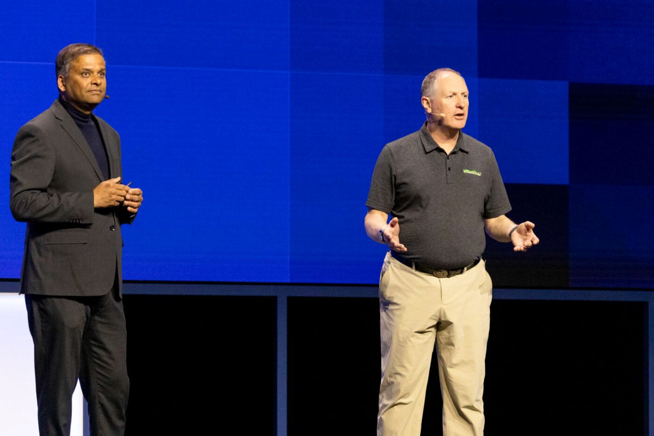 Two men are standing on stage speaking. The man on the left is wearing a dark suit, while the man on the right is in a black polo shirt and khaki pants. They appear to be engaged in a discussion or presentation against a blue geometric background.
