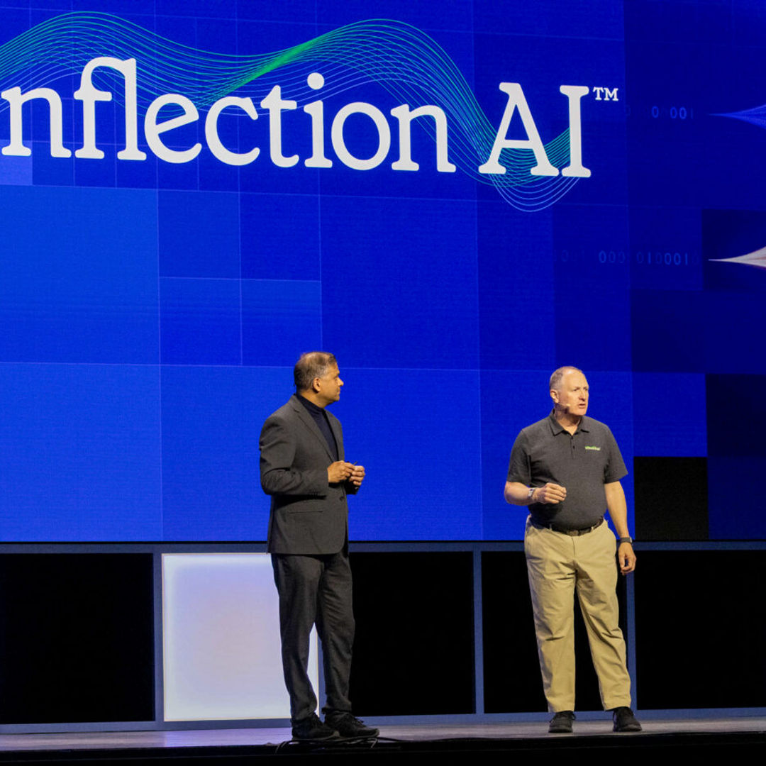 Two men stand on a stage with a large screen displaying the text Inflection AI. Colorful wave patterns are on the screen. One man wears a black suit, and the other wears a black shirt and khaki pants.