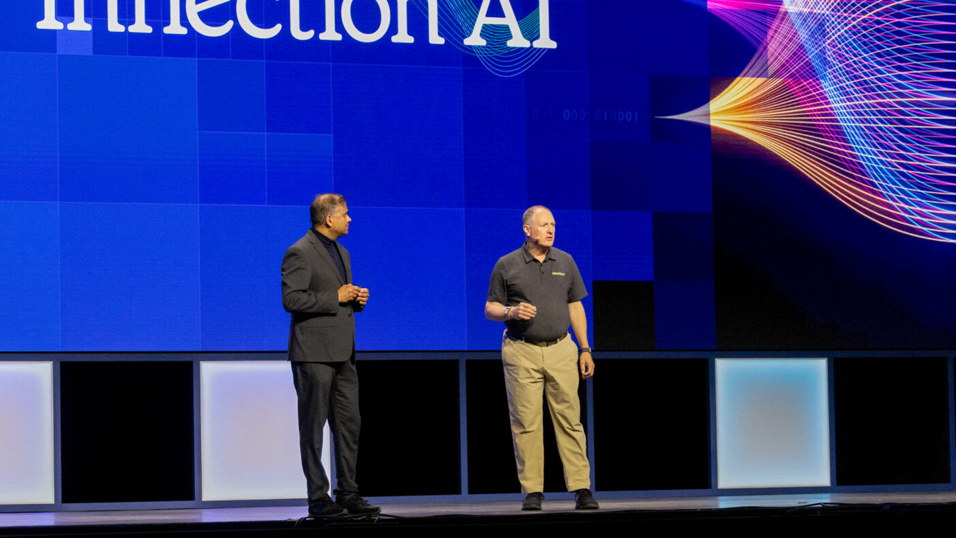 Two men stand on a stage with a large screen displaying the text Inflection AI. Colorful wave patterns are on the screen. One man wears a black suit, and the other wears a black shirt and khaki pants.