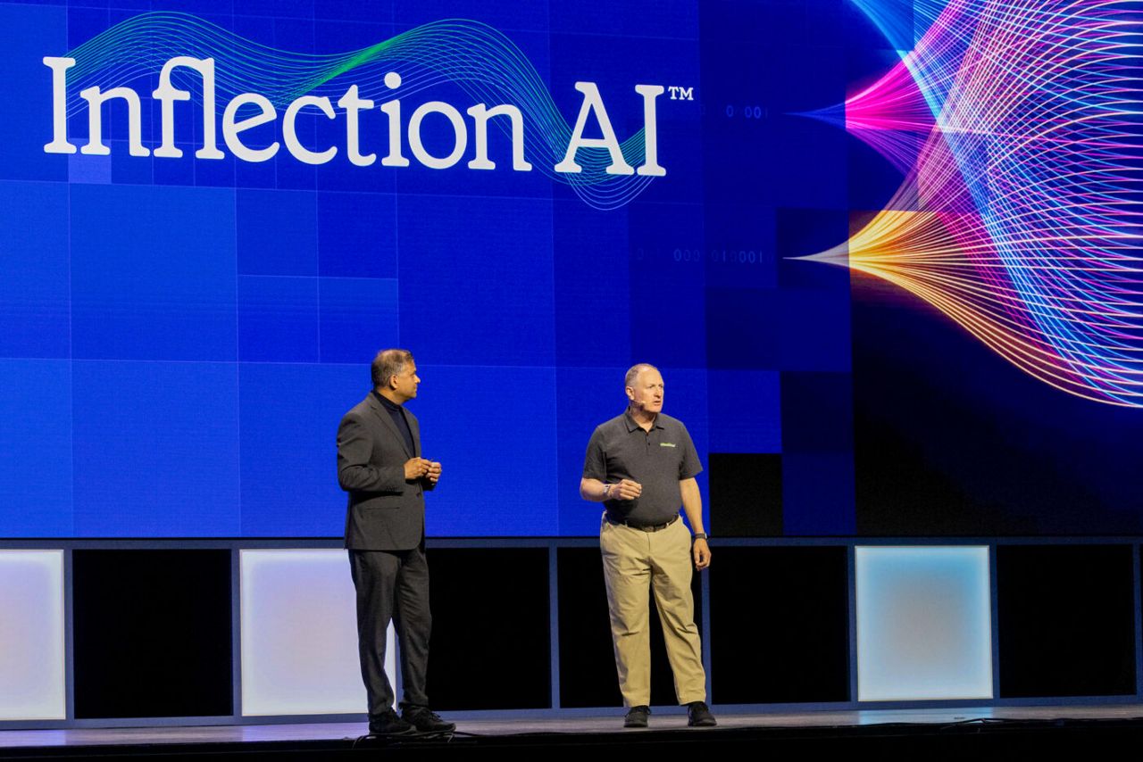 Two men stand on a stage with a large screen displaying the text Inflection AI. Colorful wave patterns are on the screen. One man wears a black suit, and the other wears a black shirt and khaki pants.