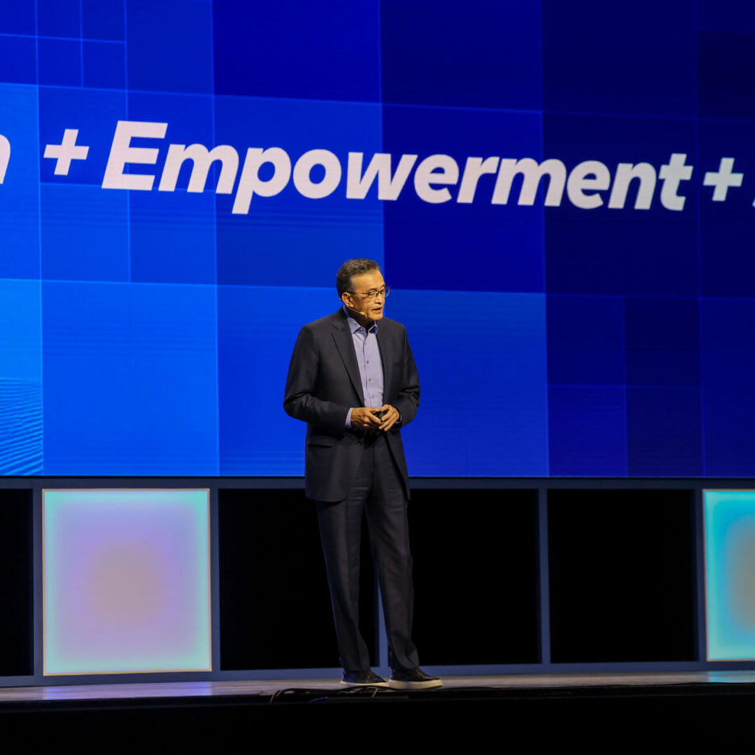 A person in a dark suit stands on a stage in front of a large blue screen with the words Innovation + Empowerment + Simplification. The stage is lit with soft, multicolored lighting from below.