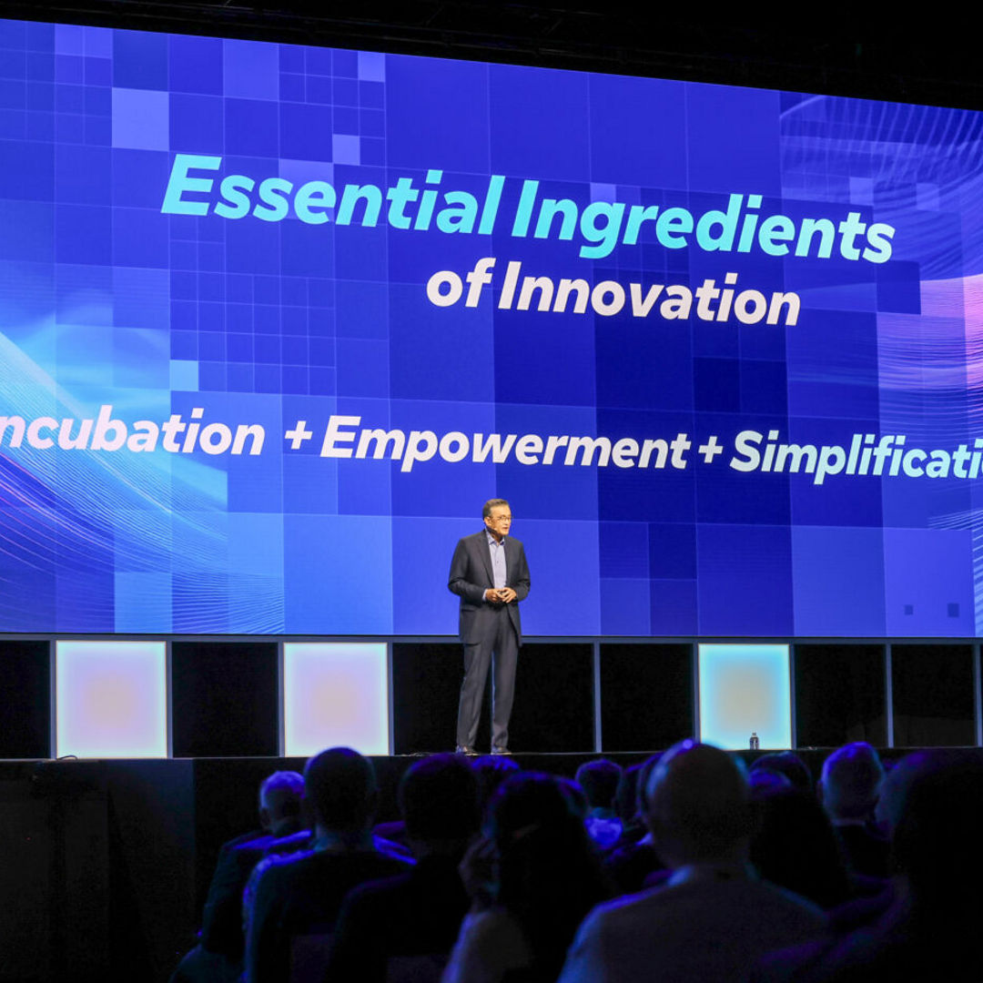 A speaker stands on a stage with a large screen behind them displaying the words Essential Ingredients of Innovation: Incubation + Empowerment + Simplification. The audience is visible in the foreground.