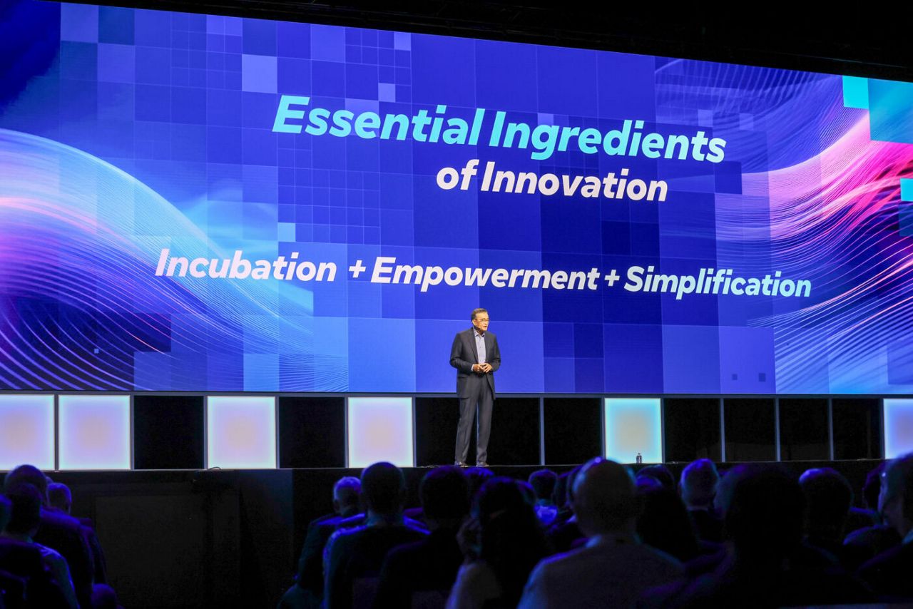 A speaker stands on a stage with a large screen behind them displaying the words Essential Ingredients of Innovation: Incubation + Empowerment + Simplification. The audience is visible in the foreground.