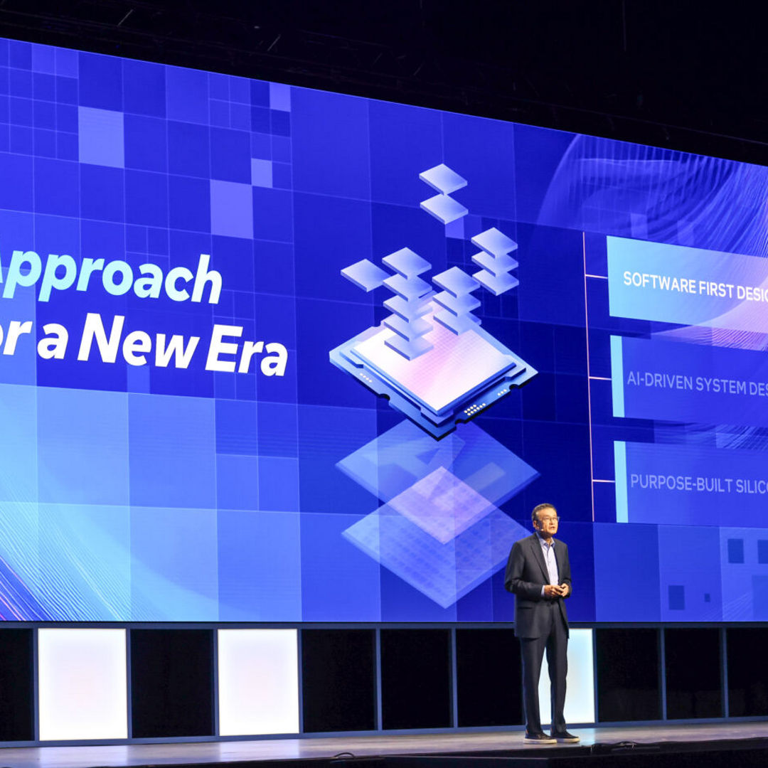 A speaker stands on stage in front of a large digital screen with the text New Approach for a New Era. The screen shows graphics of floating squares, and phrases like Software First Design Mindset and AI-Driven System Design.