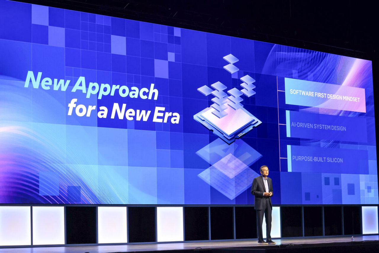 A speaker stands on stage in front of a large digital screen with the text New Approach for a New Era. The screen shows graphics of floating squares, and phrases like Software First Design Mindset and AI-Driven System Design.