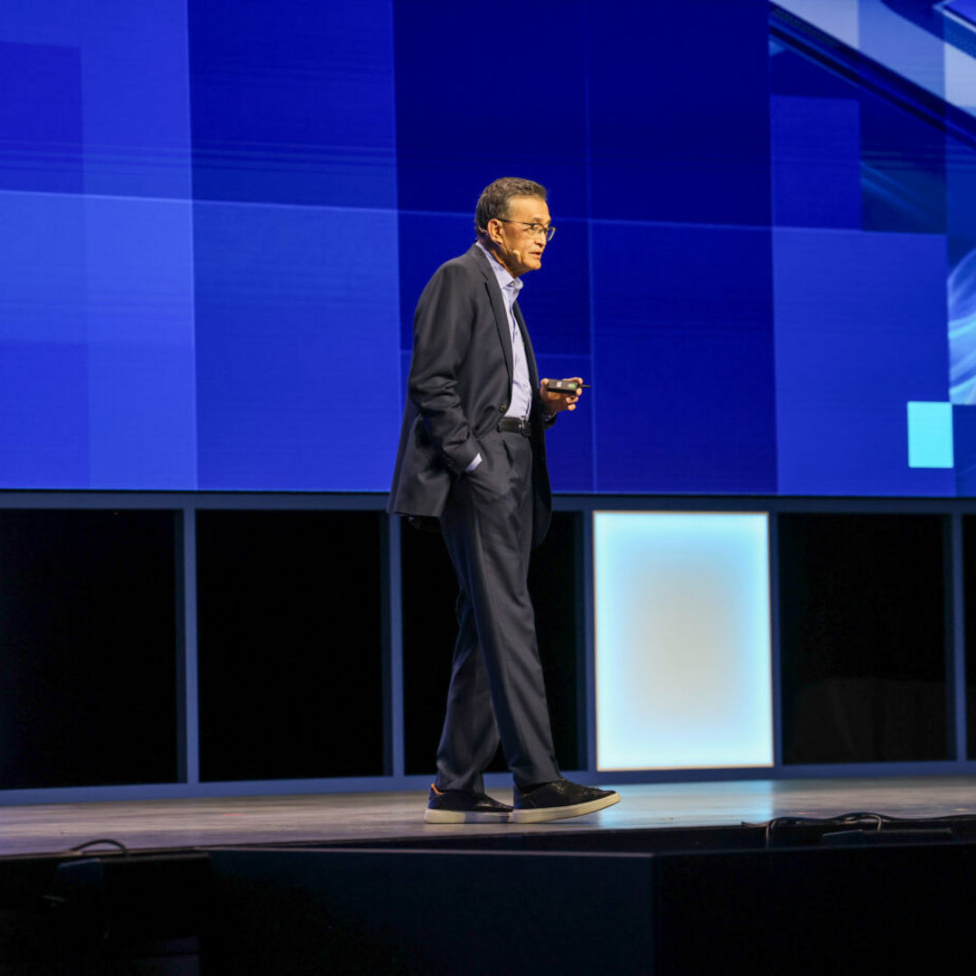 A man in a suit walks on stage during a presentation with a large blue digital screen behind him displaying abstract technology graphics. The stage floor is illuminated with light panels along the edges.