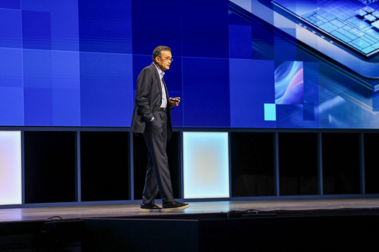 A man in a suit walks on stage during a presentation with a large blue digital screen behind him displaying abstract technology graphics. The stage floor is illuminated with light panels along the edges.