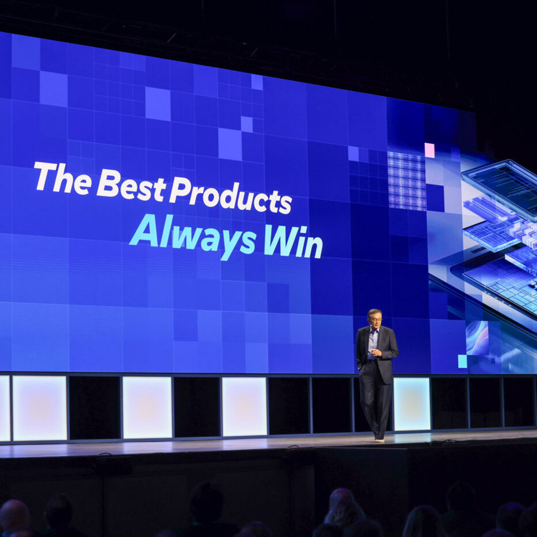 A person stands on a stage with a large blue screen behind them displaying the text The Best Products Always Win. The screen features a digital-themed graphic with circuitry and abstract patterns. An audience is visible in the foreground.