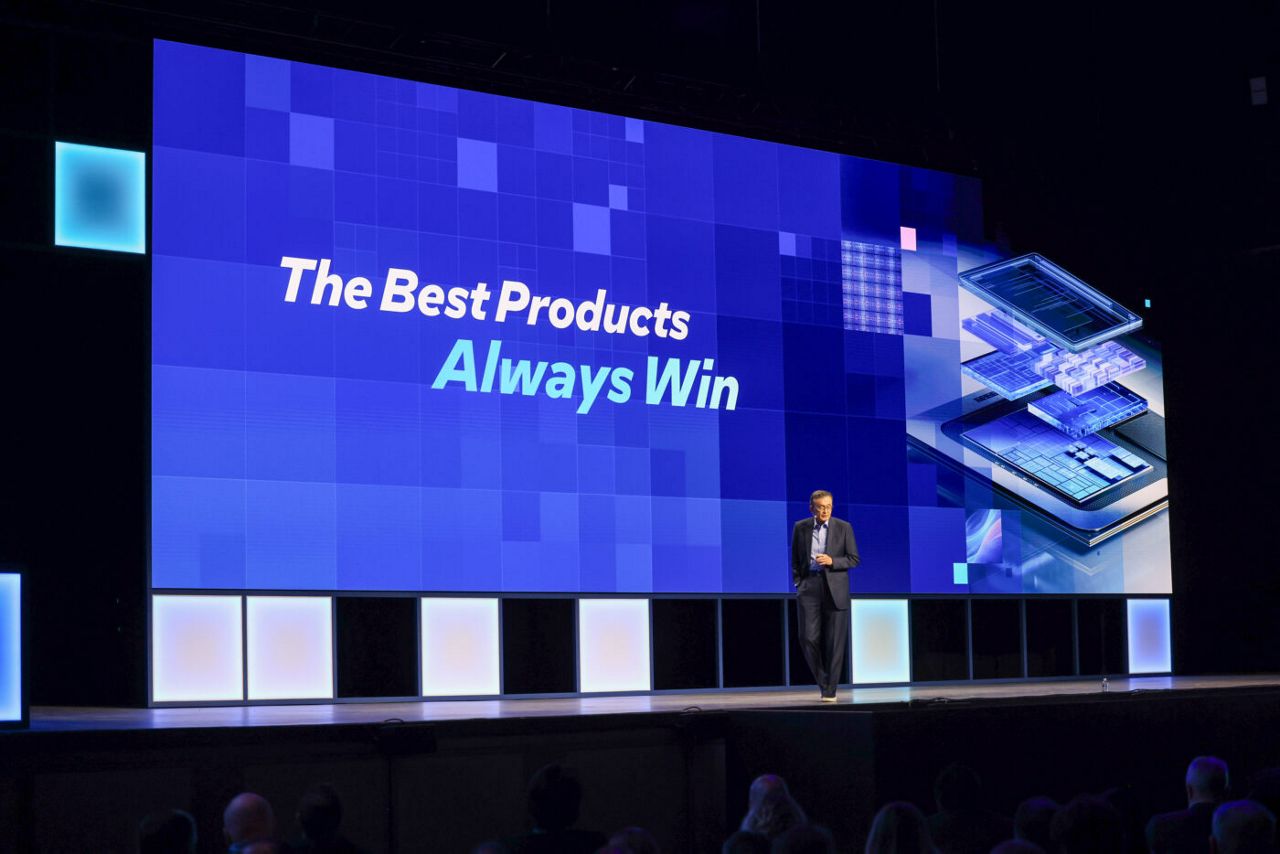 A person stands on a stage with a large blue screen behind them displaying the text The Best Products Always Win. The screen features a digital-themed graphic with circuitry and abstract patterns. An audience is visible in the foreground.