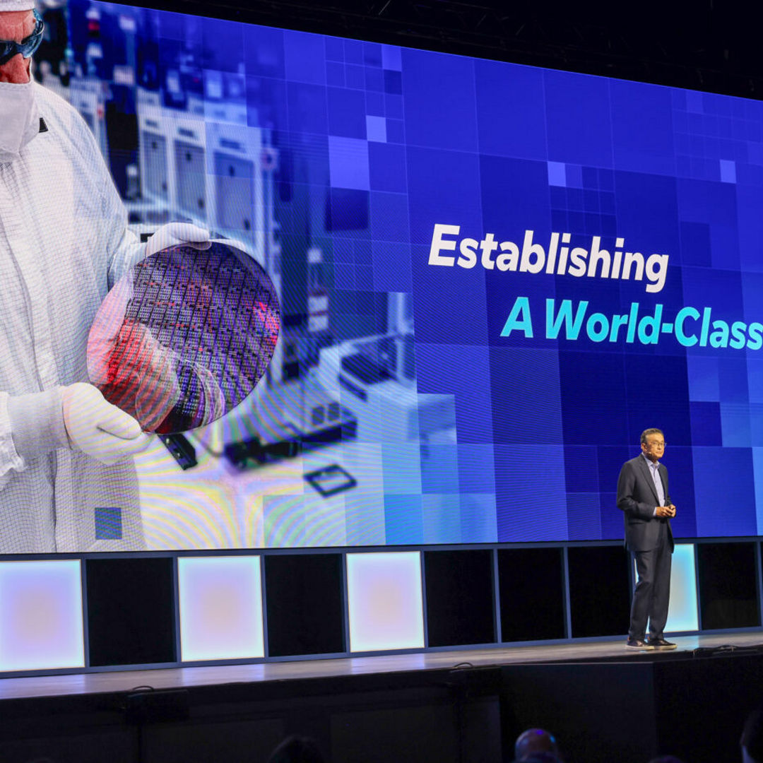 A person in a suit stands on a stage in front of a large screen displaying text that reads Establishing A World-Class Foundry. The screen also shows an image of a person holding a silicon wafer in a cleanroom setting.