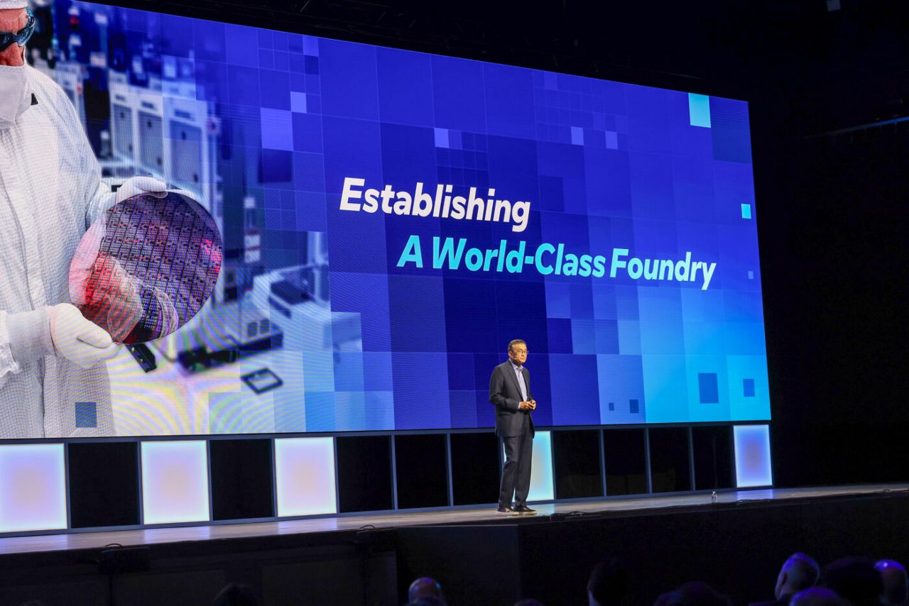 A person in a suit stands on a stage in front of a large screen displaying text that reads Establishing A World-Class Foundry. The screen also shows an image of a person holding a silicon wafer in a cleanroom setting.
