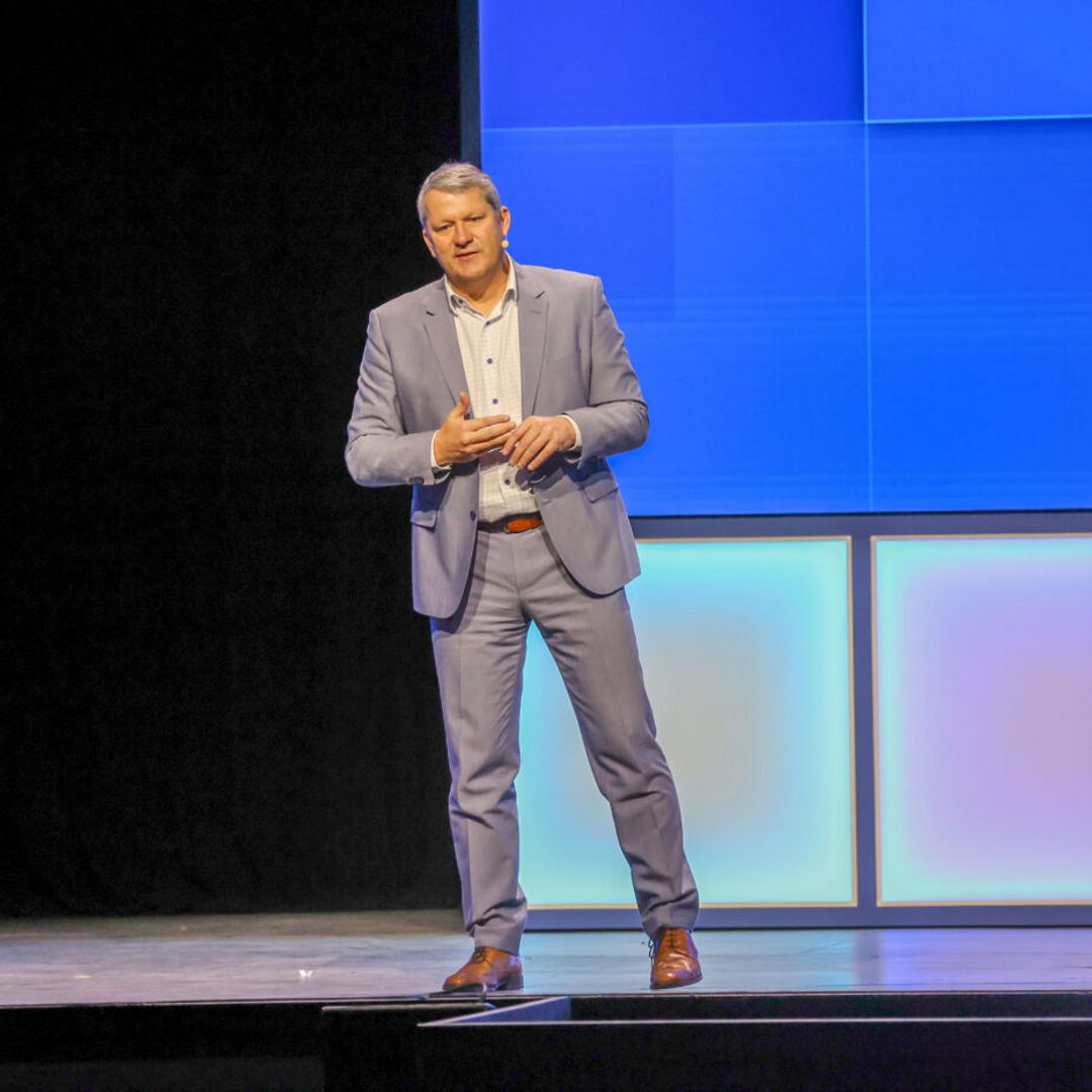 A person in a light blue suit stands on a stage with a large, brightly lit digital screen in the background. They appear to be speaking, with one hand gesturing.