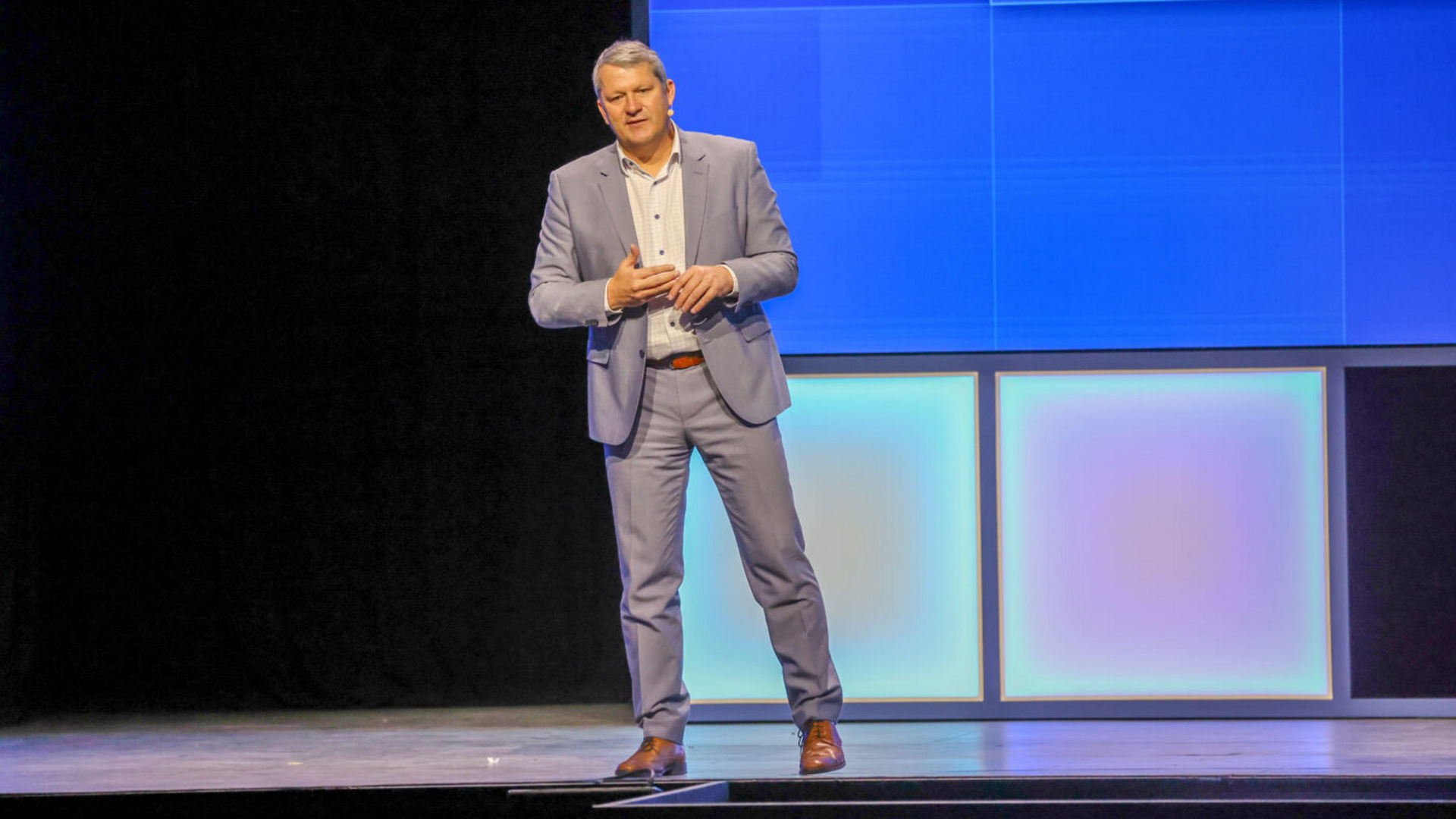 A person in a light blue suit stands on a stage with a large, brightly lit digital screen in the background. They appear to be speaking, with one hand gesturing.