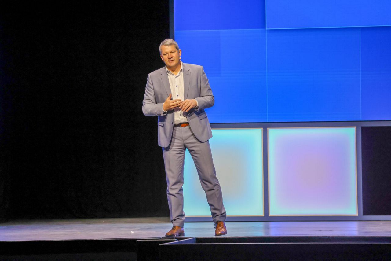 A person in a light blue suit stands on a stage with a large, brightly lit digital screen in the background. They appear to be speaking, with one hand gesturing.
