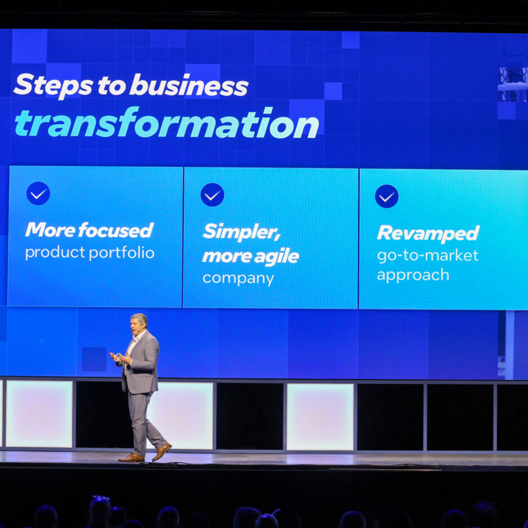 A person in business attire stands on stage, presenting a slide titled Steps to business transformation. The slide lists three points: More focused product portfolio, Simpler, more agile company, and Revamped go-to-market approach.