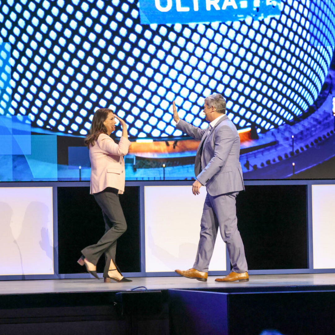 A man and a woman share a high-five on a stage, set against a backdrop of a large, pixelated screen displaying abstract graphics. The woman is in a pink blazer and black pants, and the man is in a gray suit.