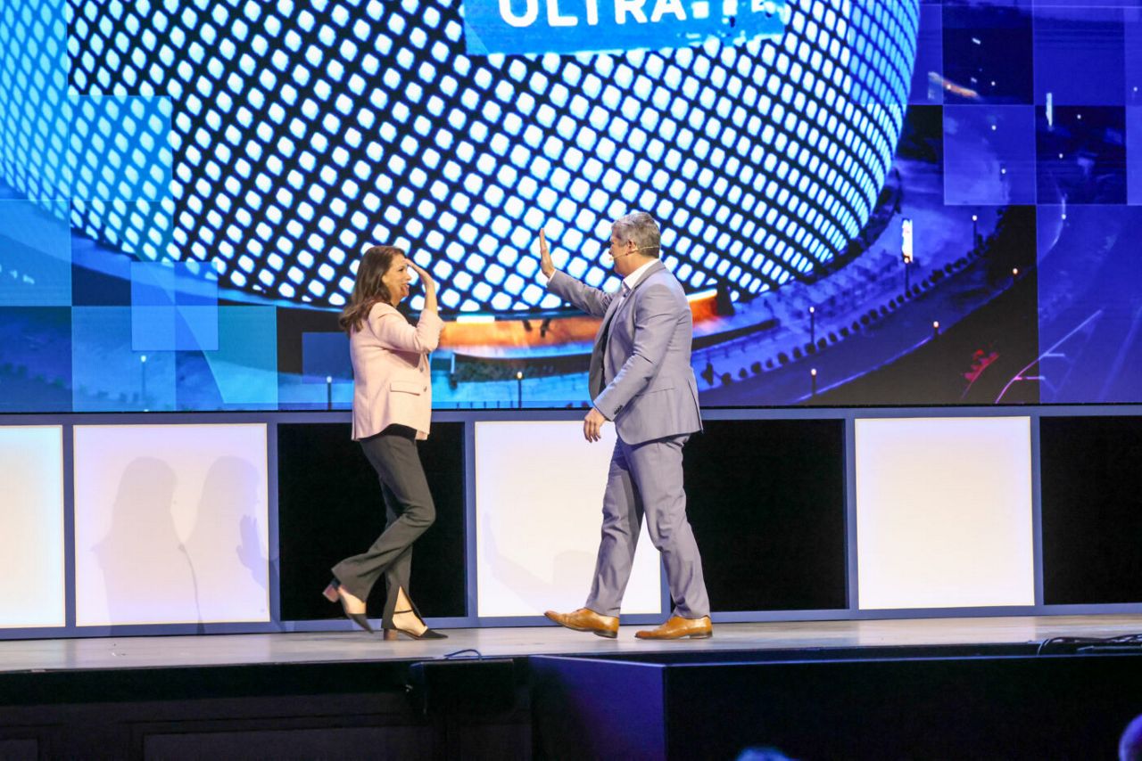 A man and a woman share a high-five on a stage, set against a backdrop of a large, pixelated screen displaying abstract graphics. The woman is in a pink blazer and black pants, and the man is in a gray suit.