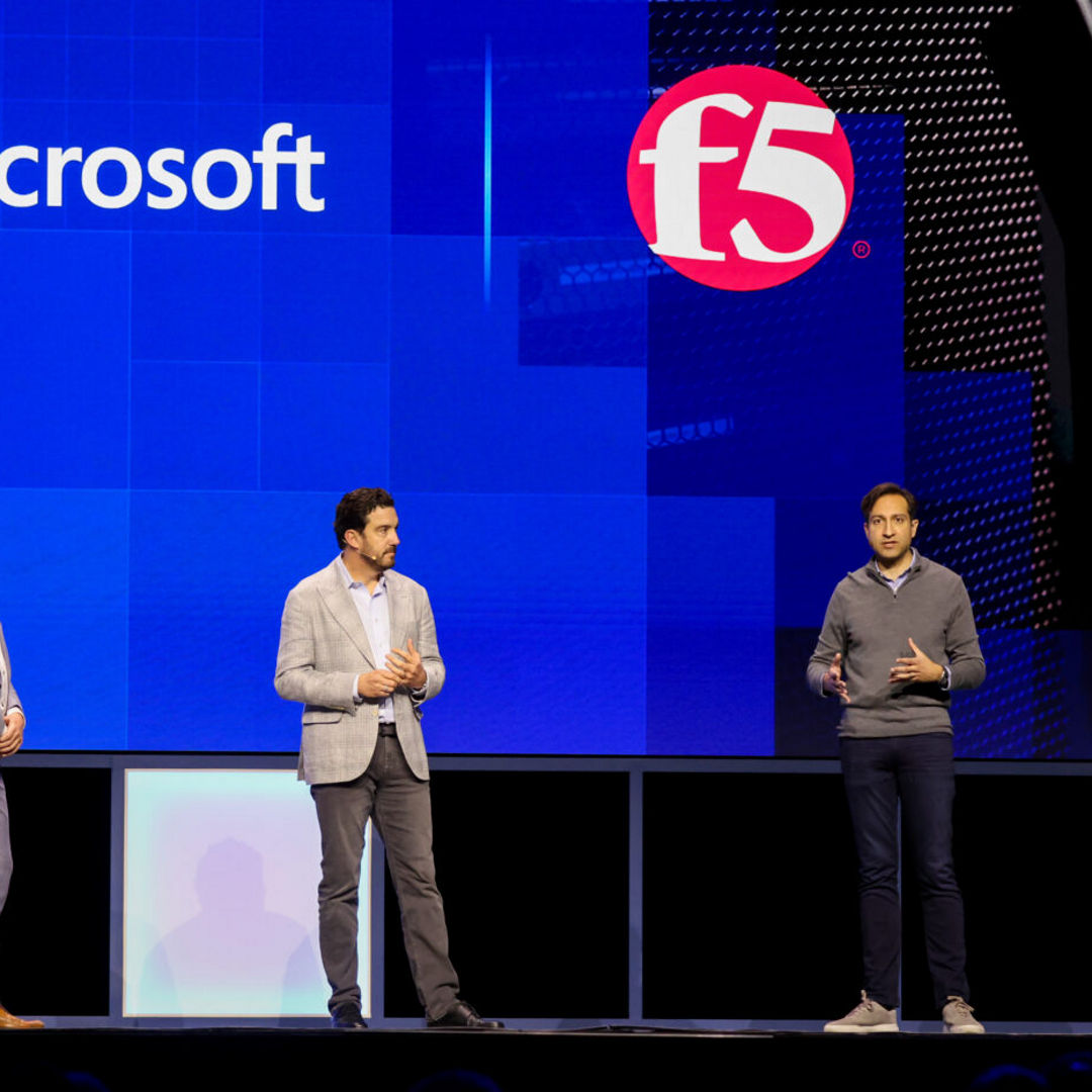 Three men stand on a stage in front of a large screen displaying Microsoft and F5 logos. The man on the right gestures with one hand while speaking. They are wearing business attire.