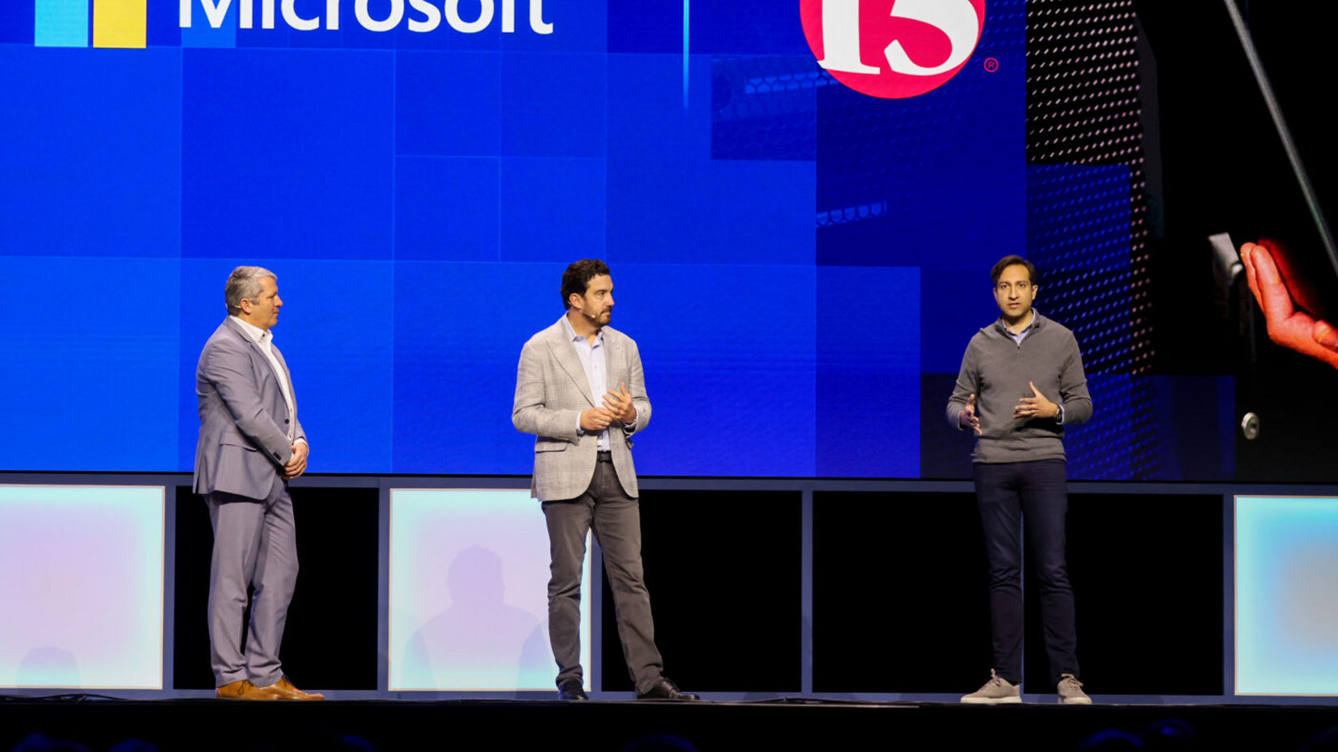 Three men stand on a stage in front of a large screen displaying Microsoft and F5 logos. The man on the right gestures with one hand while speaking. They are wearing business attire.