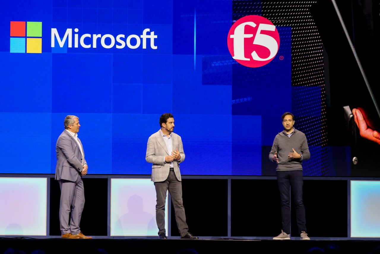 Three men stand on a stage in front of a large screen displaying Microsoft and F5 logos. The man on the right gestures with one hand while speaking. They are wearing business attire.