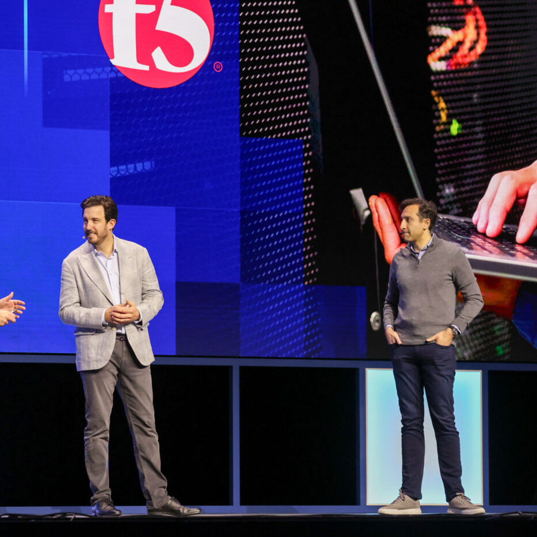 Three men are standing on a stage. Two are in conversation while another watches. A large screen behind them displays a logo and an image of hands typing on a laptop. The setting appears to be a professional event or conference.