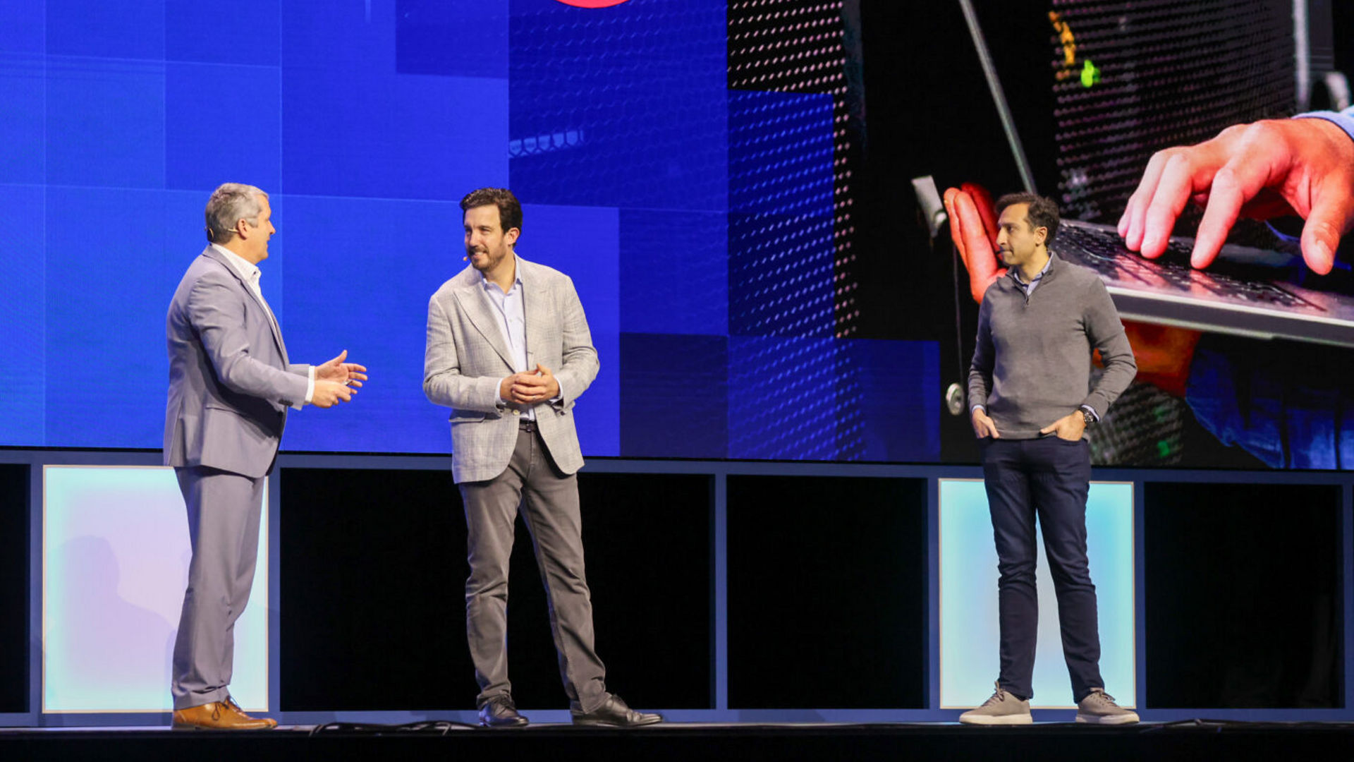 Three men are standing on a stage. Two are in conversation while another watches. A large screen behind them displays a logo and an image of hands typing on a laptop. The setting appears to be a professional event or conference.
