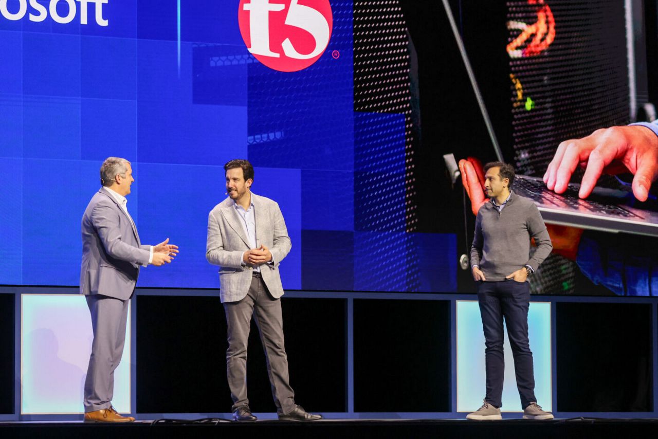 Three men are standing on a stage. Two are in conversation while another watches. A large screen behind them displays a logo and an image of hands typing on a laptop. The setting appears to be a professional event or conference.