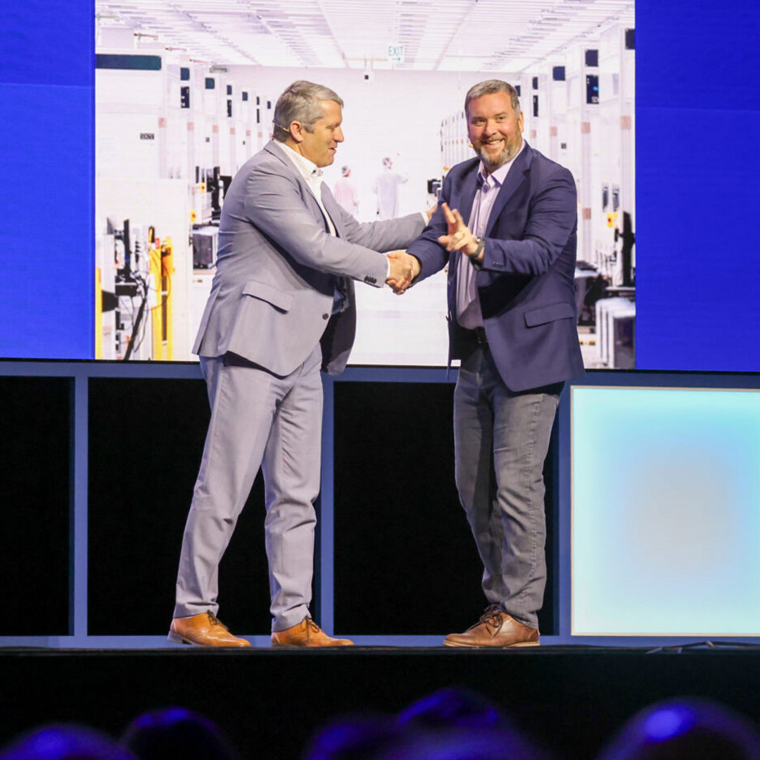 Two men in suits are shaking hands on a stage, smiling at each other. They stand in front of large screens displaying images of a factory setting. The stage is lit with blue lighting.