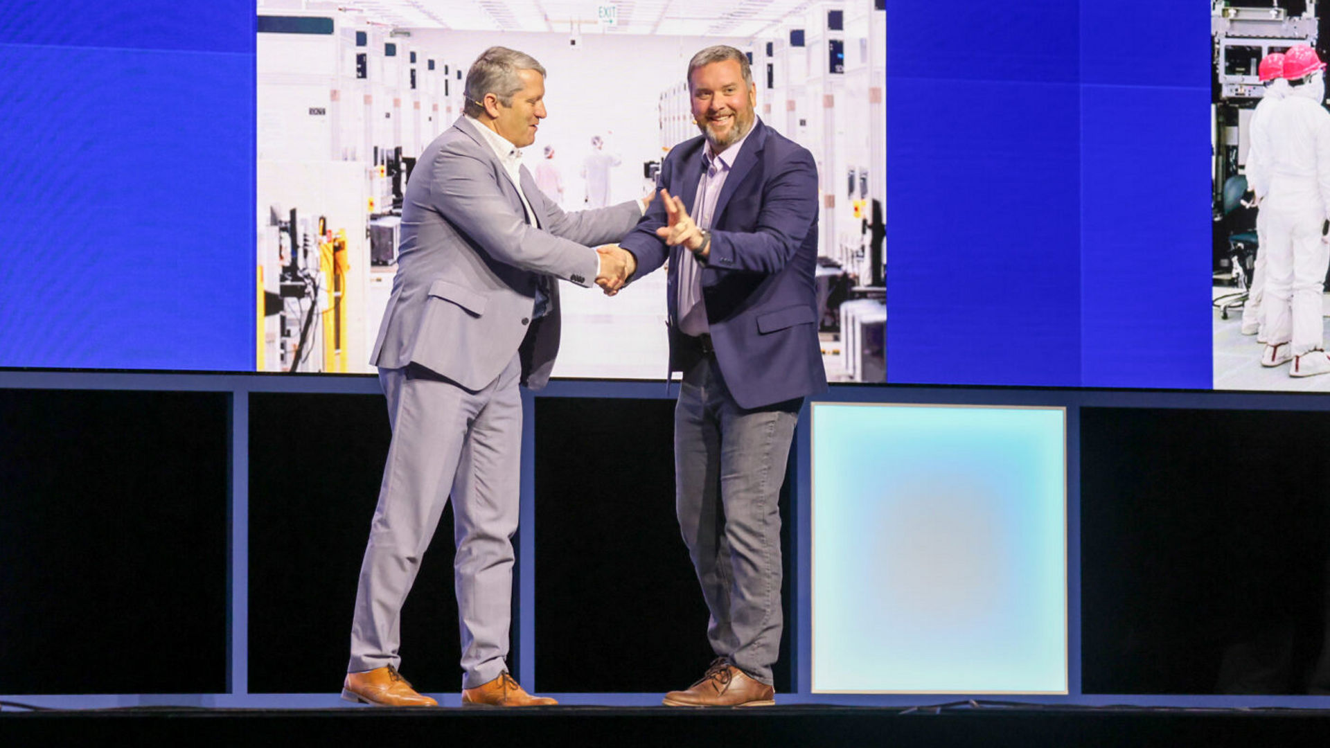 Two men in suits are shaking hands on a stage, smiling at each other. They stand in front of large screens displaying images of a factory setting. The stage is lit with blue lighting.