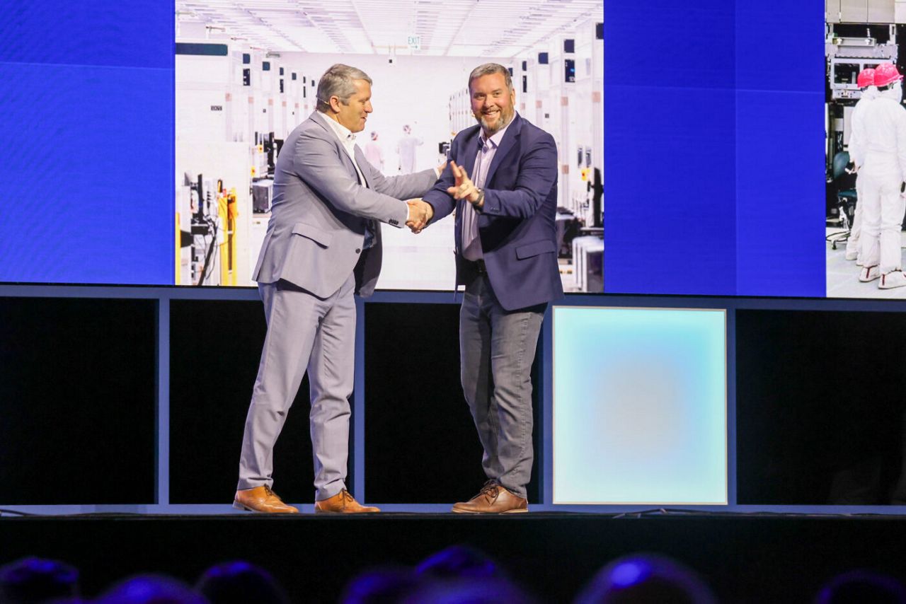 Two men in suits are shaking hands on a stage, smiling at each other. They stand in front of large screens displaying images of a factory setting. The stage is lit with blue lighting.
