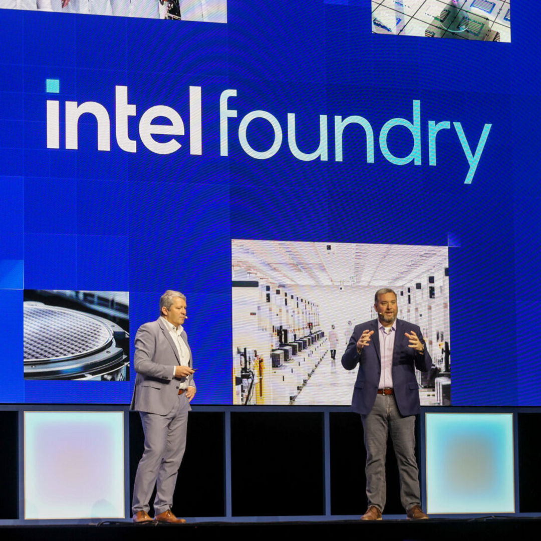 Two men stand on a stage in front of a large screen displaying Intel Foundry. The background features images of manufacturing facilities. Both are dressed in suits, speaking and gesturing as if presenting to an audience.