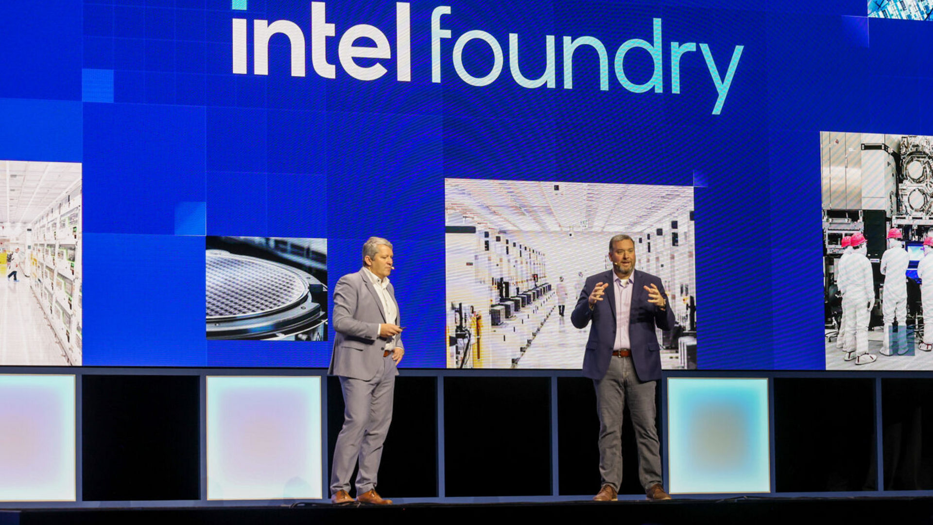Two men stand on a stage in front of a large screen displaying Intel Foundry. The background features images of manufacturing facilities. Both are dressed in suits, speaking and gesturing as if presenting to an audience.