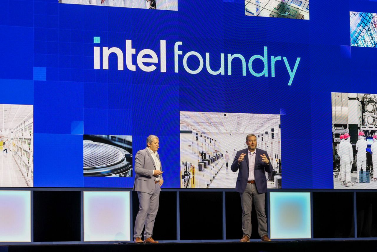 Two men stand on a stage in front of a large screen displaying Intel Foundry. The background features images of manufacturing facilities. Both are dressed in suits, speaking and gesturing as if presenting to an audience.
