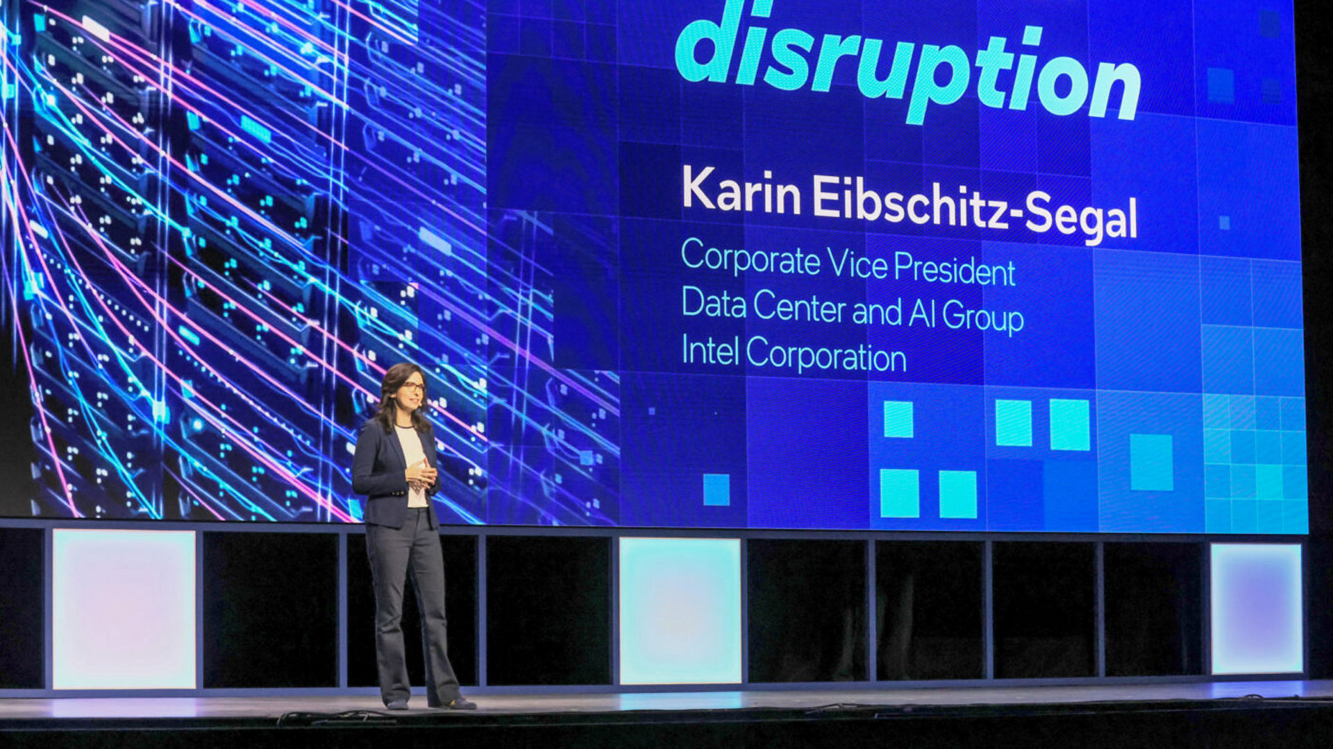 A person is standing on a stage presenting. The large screen behind them displays the text Data center disruption and Karin Eibschitz-Segal, Corporate Vice President, Data Center and AI Group, Intel Corporation.