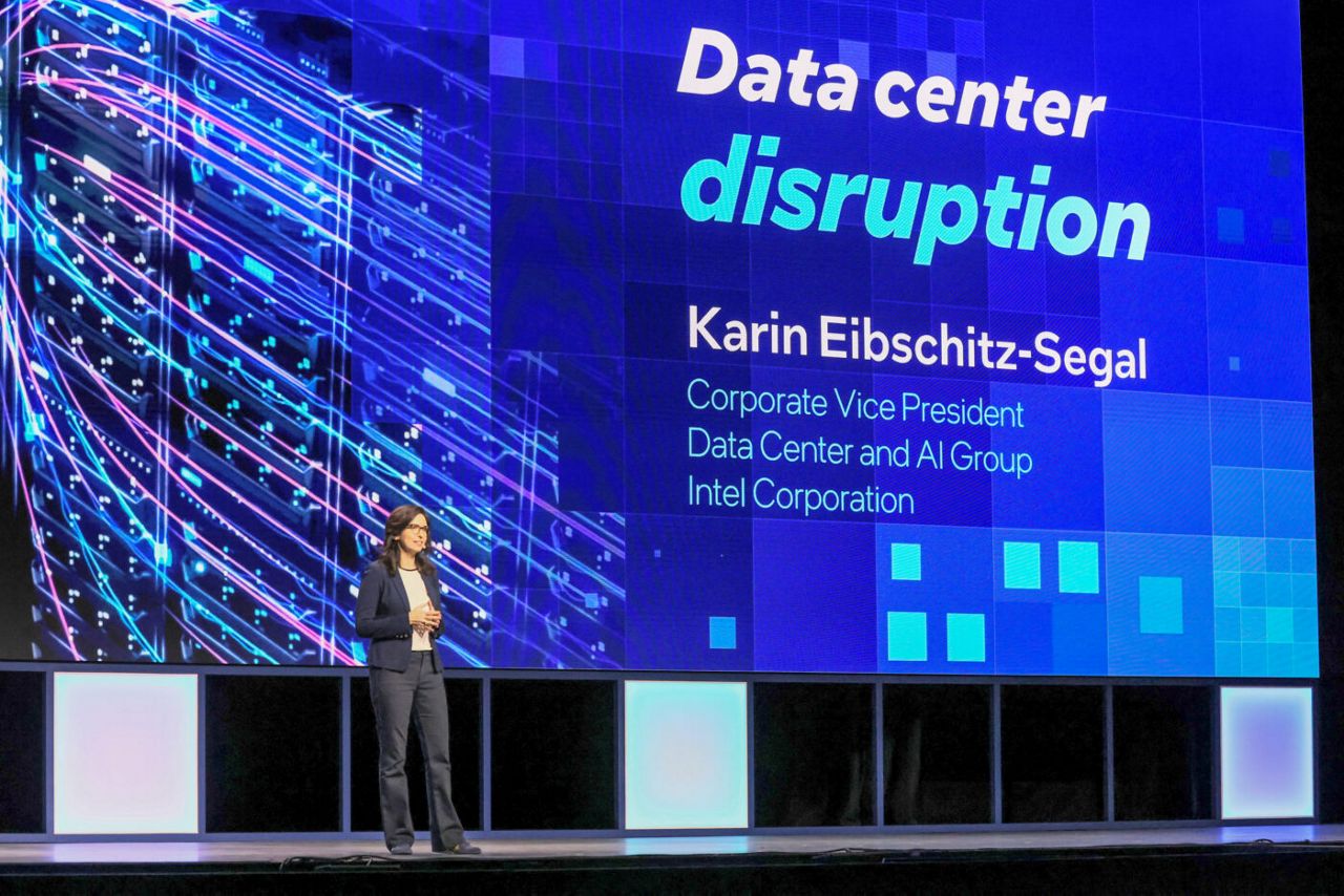 A person is standing on a stage presenting. The large screen behind them displays the text Data center disruption and Karin Eibschitz-Segal, Corporate Vice President, Data Center and AI Group, Intel Corporation.