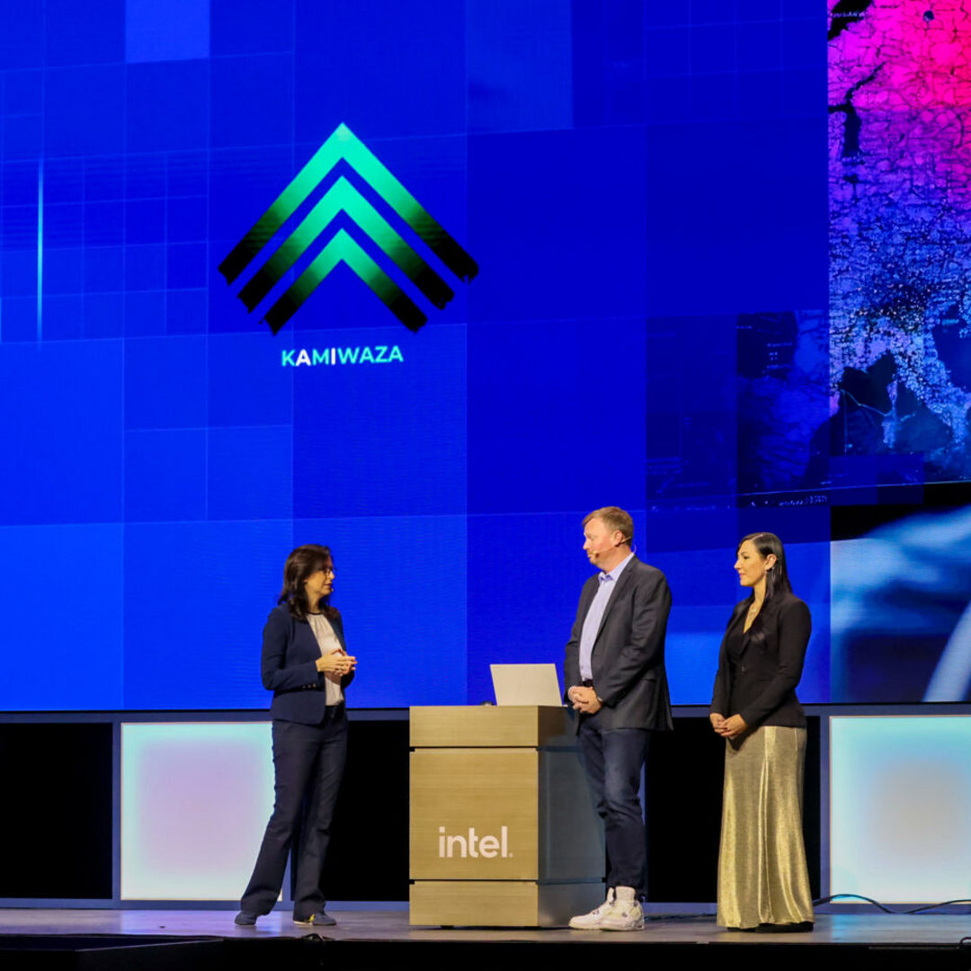 Three individuals stand on a stage in front of screens displaying the Intel logo and another logo labeled KAMIWAZA. One person is behind a podium with a laptop. The setting appears to be a professional or tech event.